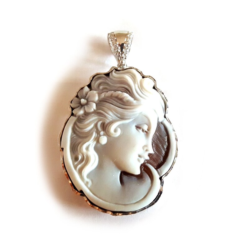May include: A cameo pendant featuring a carved portrait of a woman with a flower in her hair. The cameo is set in a silver-tone frame with a scalloped edge.