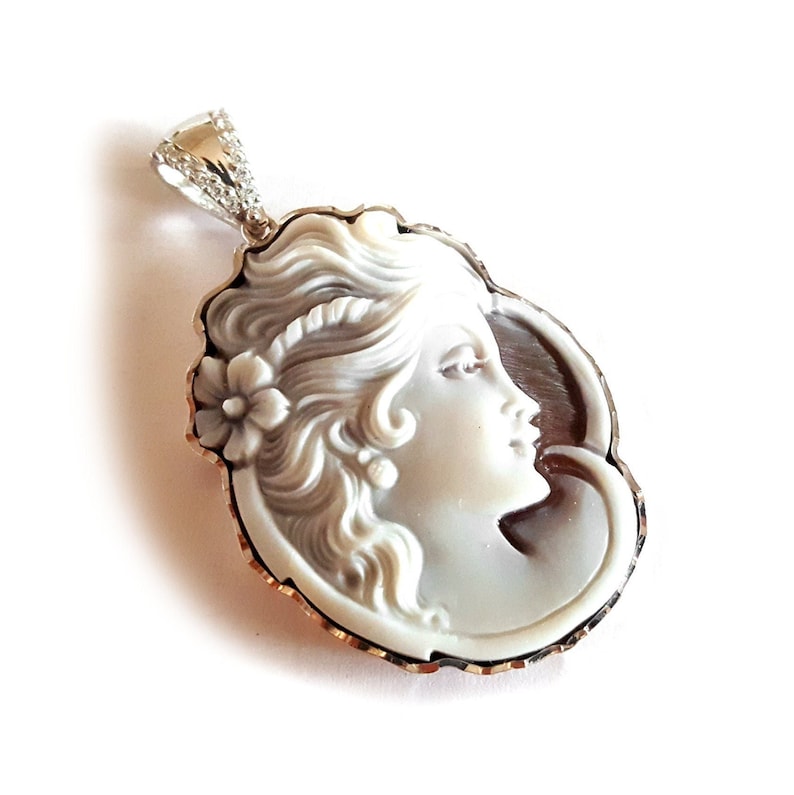 May include: A cameo pendant featuring a carved portrait of a woman with detailed hair and a flower. The cameo is set in a silver frame with a scalloped edge.