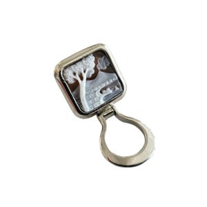 May include: A silver key chain with a square cameo featuring a white cameo of a tree and a brown background.