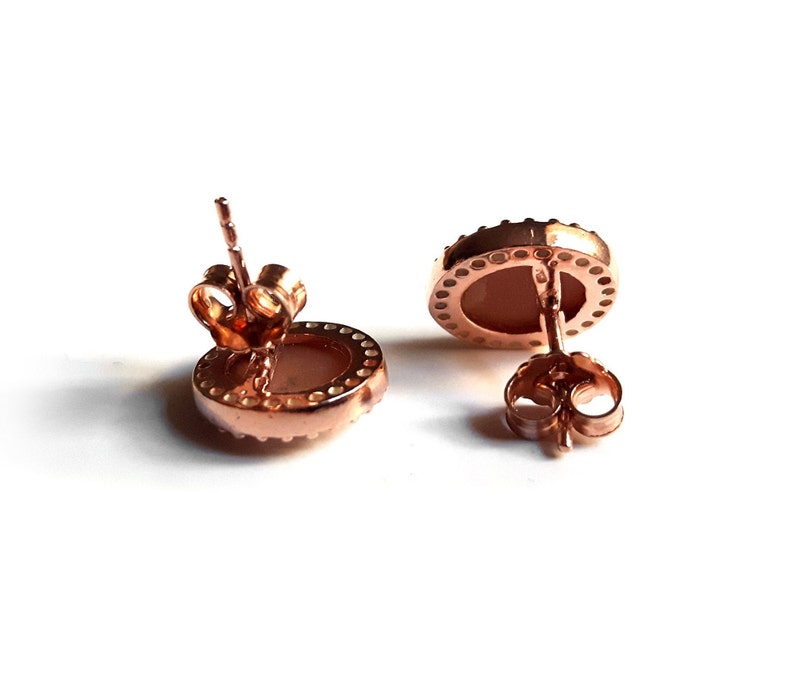 May include: A pair of rose gold stud earrings with a round, white stone set in a bezel setting. The earrings have a post and butterfly back.