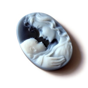 May include: A white cameo carving of a mother and child, with the mother's face and hair carved in detail. The cameo is set in a black oval-shaped base.