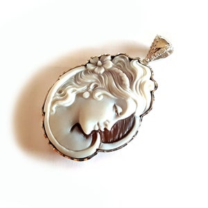 May include: A white cameo pendant with a detailed carving of a woman's face, set in a silver frame with a decorative edge. The pendant is attached to a silver chain with a small, clear crystal.