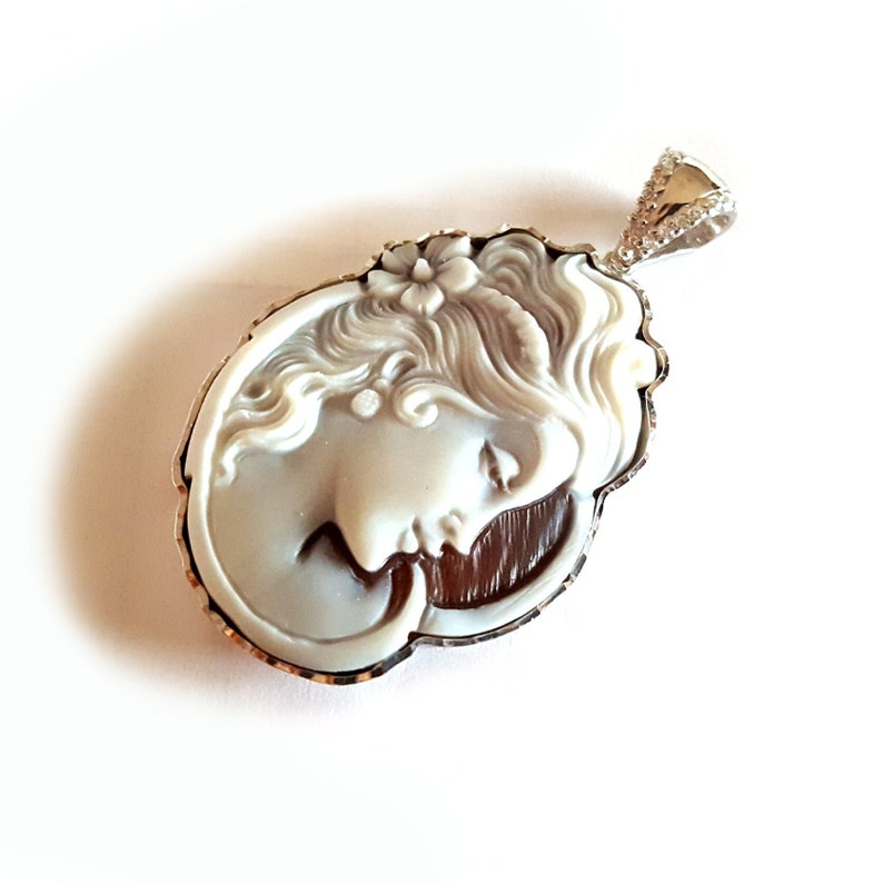May include: A white cameo pendant with a detailed carving of a woman's face, set in a silver frame with a decorative edge. The pendant is attached to a silver chain with a small, clear crystal.