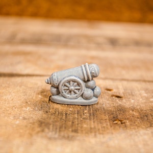 May include: A small, gray ceramic figurine of a cannon with wheels and cannonballs. The cannon is on a wooden surface.