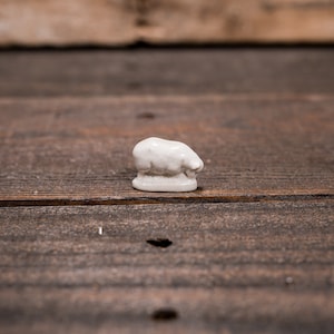 May include: A small, white ceramic chef's hat figurine. The hat is detailed with a rounded top and a brim, set against a rustic, dark wood background. The figurine is a miniature representation of a chef's hat.