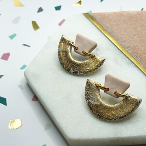 May include: A pair of gold and pink ceramic earrings with a geometric design. The earrings are on a white and pink marble surface.