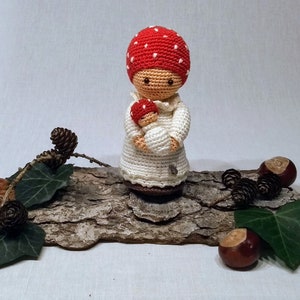Puede incluir: Muñeca de ganchillo hecha a mano, con un gorro de seta rojo con lunares blancos y un vestido blanco, sosteniendo una seta más pequeña. La muñeca se exhibe sobre un trozo de madera con hojas, piñas y castañas.