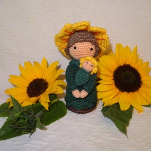May include: A crocheted doll with brown hair and a yellow sunflower hat holding a smaller crocheted doll. The dolls are surrounded by sunflowers.