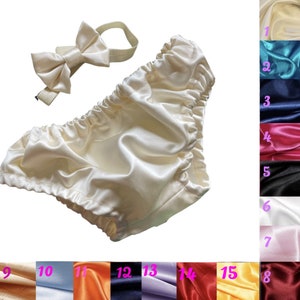 May include: A set of ivory satin baby bloomers with a matching ivory satin bow tie headband. The bloomers have a gathered elastic waistband and are made of soft, luxurious satin. The headband is adjustable and has a bow on top.
