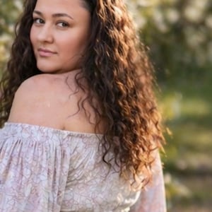 May include: A woman with long, curly brown hair looks over her shoulder. She wears an off-the-shoulder top with a floral pattern in shades of pink and white. The background is blurred with greenery.