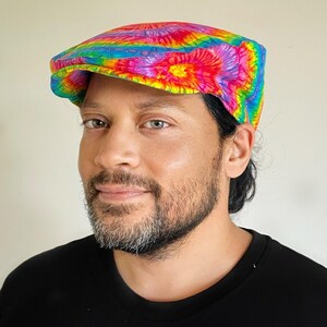 Peut inclure: Casquette plate tie-dye aux couleurs de l'arc-en-ciel. La casquette est portée par une personne portant un haut noir. Le texte "Bazzybears" est dans le coin supérieur gauche.