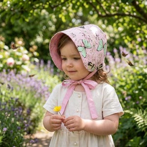 Peut inclure: Une jeune enfant porte un bonnet rose orné de papillons et un nœud assorti. L'enfant tient une fleur jaune dans un jardin, entouré de papillons et de fleurs.