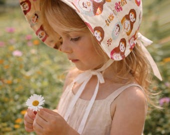 Könnte beinhalten: Ein beiger Sonnenhut mit Krempe, verziert mit bunten Matrjoschka-Puppen und floralen Mustern. Der Hut ist mit weißen Bändern gebunden. Das Kind trägt ein beiges Kleid und hält eine Gänseblümchen in einem Blumenfeld.
