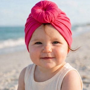 Peut inclure: Un turban rose vif pour bébé, avec un nœud sur le dessus. L'image montre un bébé souriant portant le chapeau sur une plage, avec la mer et le ciel en arrière-plan. Le bébé porte un débardeur blanc.