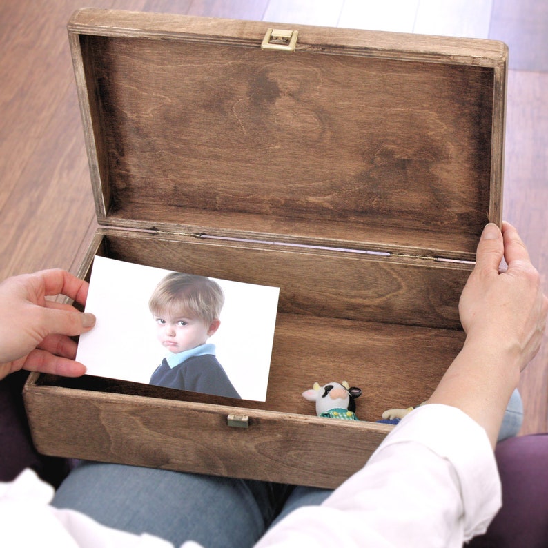 K&ouml;nnte beinhalten: Eine Holzkiste mit einem Scharnierdeckel ist ge&ouml;ffnet und zeigt ein Foto eines jungen Jungen und eine kleine Spielzeugkuh im Inneren. Die Kiste ist aus dunkelbraunem Holz und hat einen Messingverschluss.