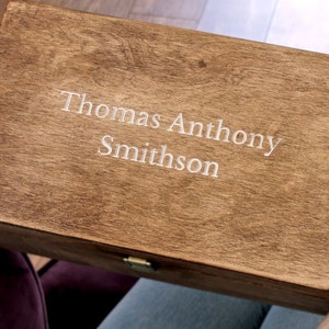 K&ouml;nnte beinhalten: Eine Holzkiste mit einem Scharnierdeckel und einem Messingverschluss. Die Kiste ist mit dem Namen "Thomas Anthony Smithson" graviert.