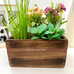 May include: A dark brown wooden planter box containing green grass, pink and yellow flowers, and green leafy plants. The box has a copper-coloured band with the text "LOVE YOU LONG THYME". The planter is a decorative item.