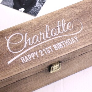 Puede incluir: Una caja de madera con tapa con bisagras y cierre de lat&oacute;n. La caja est&aacute; grabada con el texto "Charlotte Happy 21st Birthday" en una fuente cursiva.