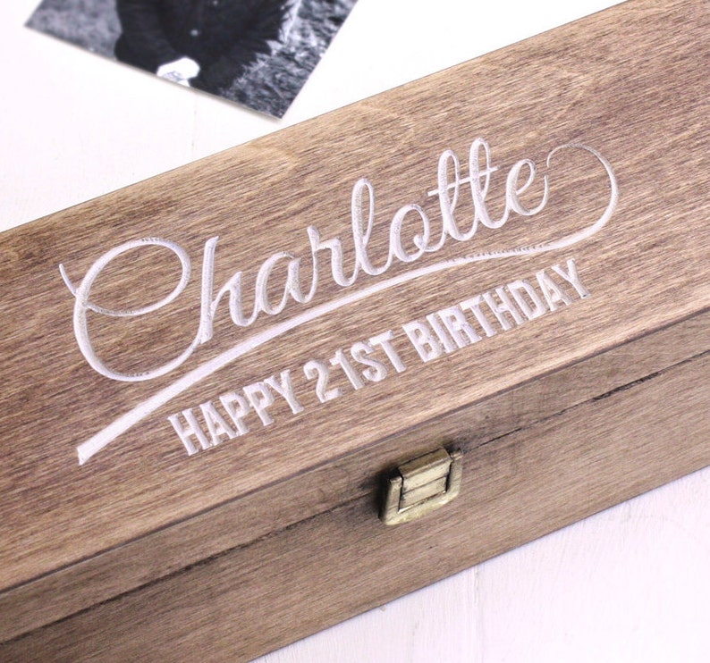 Puede incluir: Una caja de madera con tapa con bisagras y cierre de lat&oacute;n. La caja est&aacute; grabada con el texto "Charlotte Happy 21st Birthday" en una fuente cursiva.