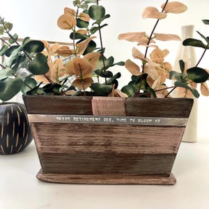 May include: A wooden planter with a silver band engraved with the text "Happy Retirement Dee, Time to Bloom xx". The planter is filled with a leafy green plant with pink leaves.