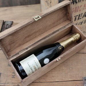 Puede incluir: Una caja de madera con tapa abatible, abierta para revelar una botella de champ&aacute;n. La botella est&aacute; etiquetada con "Louis Delaunay" y "Champagne".