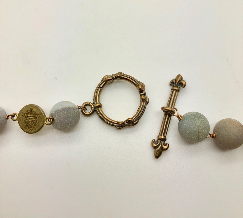 K&ouml;nnte beinhalten: Nahaufnahme einer Halskette mit runden, grauen Perlen und antiken Messingfarbenen Akzenten. Die Halskette hat einen Knebelverschluss mit Fleur-de-Lis-Design. Ein kleiner, runder Messinganh&auml;nger mit Wappen ist ebenfalls sichtbar.