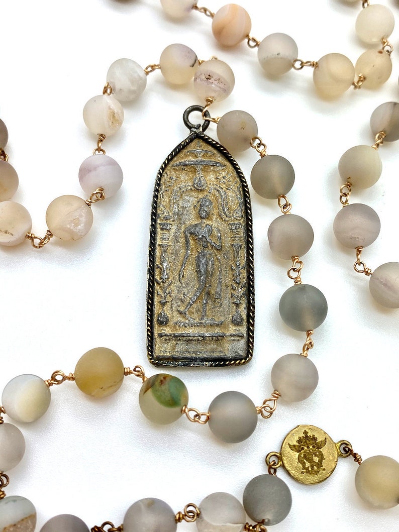 May include: A gray and beige beaded necklace with a gold chain and a silver pendant featuring a detailed image of a standing figure. The pendant is rectangular with a pointed top and bottom.