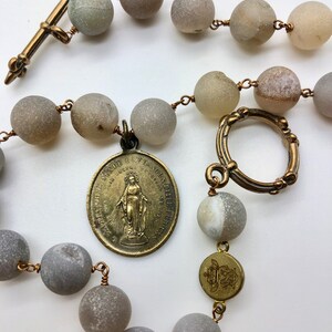 K&ouml;nnte beinhalten: Ein Rosenkranz mit gro&szlig;en, runden, grauen Perlen, die auf Kupferdraht aufgef&auml;delt sind. Er hat ein bronzefarbenes Kreuz, einen kreisf&ouml;rmigen Verschluss und ein Medaillon mit einer religi&ouml;sen Figur. Die Perlen haben ein mattes Finish.