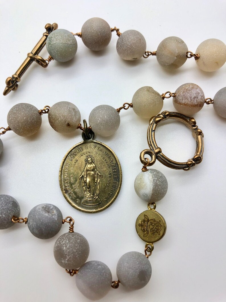 K&ouml;nnte beinhalten: Ein Rosenkranz mit gro&szlig;en, runden, grauen Perlen, die auf Kupferdraht aufgef&auml;delt sind. Er hat ein bronzefarbenes Kreuz, einen kreisf&ouml;rmigen Verschluss und ein Medaillon mit einer religi&ouml;sen Figur. Die Perlen haben ein mattes Finish.