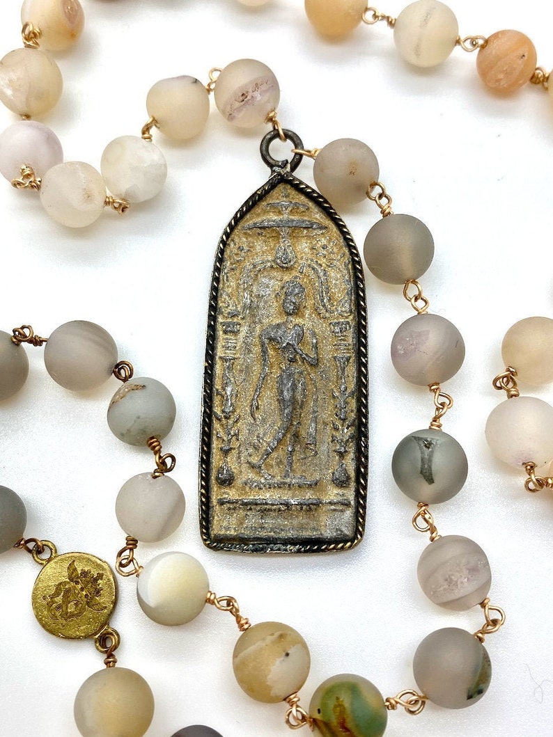 May include: A long necklace made of light brown and gray agate beads with a gold metal chain. The necklace has a large, rectangular pendant with a detailed image of a person in a standing pose. The pendant is made of metal with a dark patina.