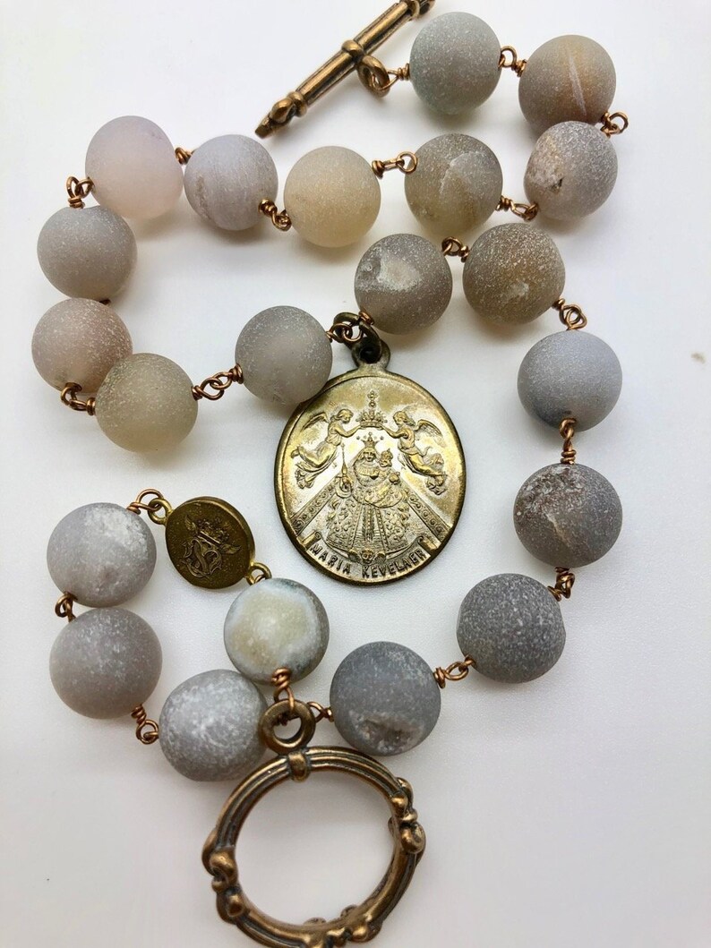 K&ouml;nnte beinhalten: Ein Rosenkranz mit matten grauen Perlen und Kupferdrahtverbindungen. Der Rosenkranz hat einen dekorativen Metallanh&auml;nger mit dem Text "MARIA REVELADORA" und einen kreuzf&ouml;rmigen Verschluss. Die Perlen haben einen Durchmesser von etwa 1,3 cm.