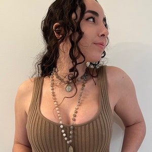 May include: A woman wearing a brown ribbed tank top with two necklaces. The necklaces are made of beads and have a pendant on each. The pendant on the longer necklace is a silver charm with a figure on it. The pendant on the shorter necklace is a silver coin.