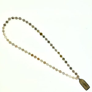 May include: A long necklace made of light brown and gray round beads with a small rectangular pendant at the end. The pendant has a design of a person on it.
