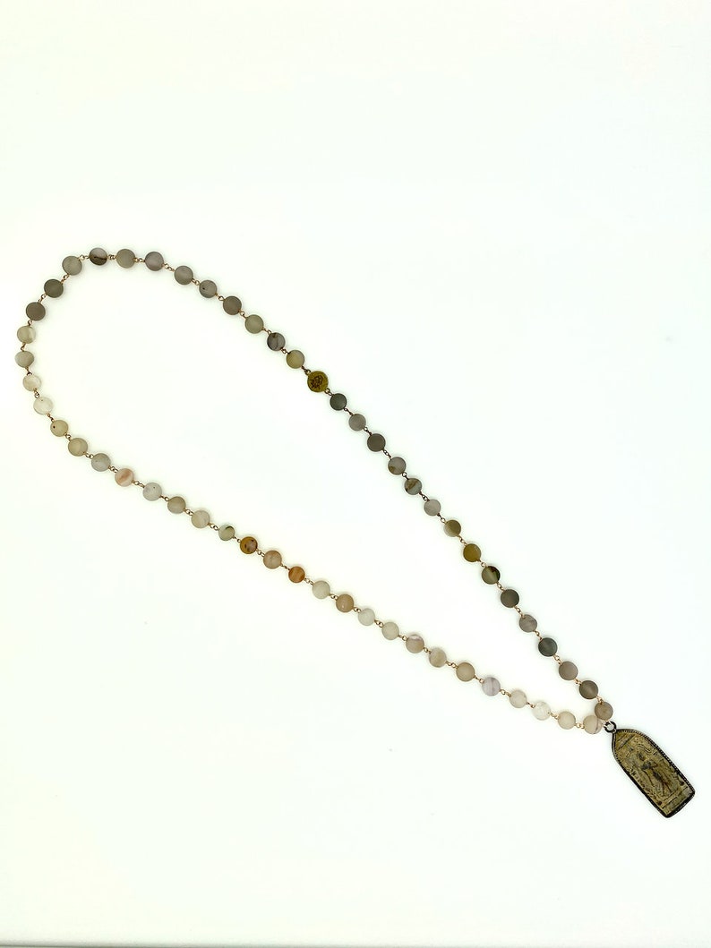 May include: A long necklace made of light brown and gray round beads with a small rectangular pendant at the end. The pendant has a design of a person on it.