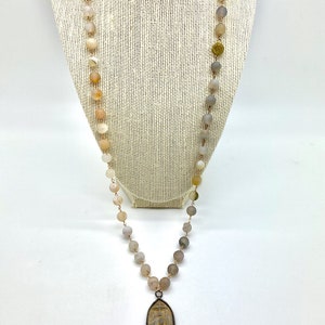 May include: A long necklace with a pendant featuring a detailed carving. The necklace is made of light brown and gray beads.
