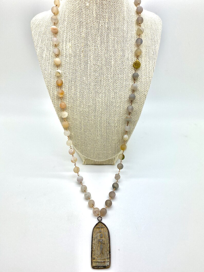 May include: A long necklace with a pendant featuring a detailed carving. The necklace is made of light brown and gray beads.