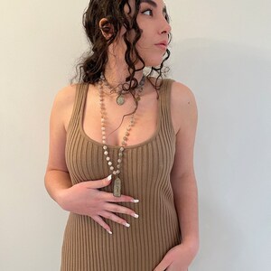 K&ouml;nnte beinhalten: Eine Frau tr&auml;gt ein geripptes Tankkleid in Beige und eine lange Perlenkette mit Anh&auml;nger. Die Halskette hat verschiedene Perlen und einen rechteckigen Anh&auml;nger. Das Kleid ist einfarbig und hat einen Rundhalsausschnitt.