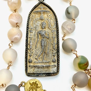 May include: A long necklace with a gold chain and grey agate beads. The necklace has a large pendant with a silver and gold design of a standing figure. The pendant is attached to the chain with a gold ring.