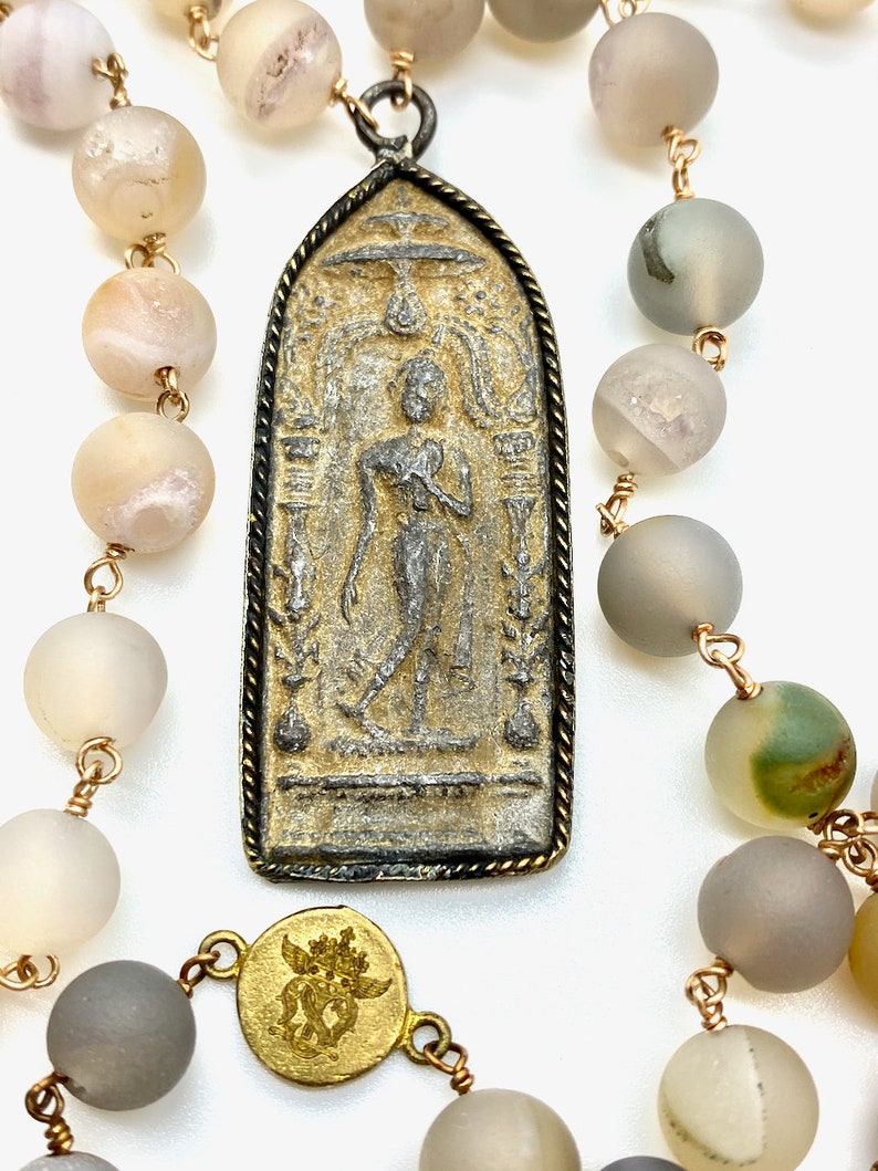 May include: A long necklace with a gold chain and grey agate beads. The necklace has a large pendant with a silver and gold design of a standing figure. The pendant is attached to the chain with a gold ring.