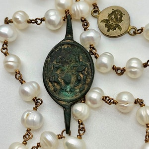 May include: A white pearl necklace with a bronze pendant featuring a detailed design. The necklace is strung on a gold chain and includes a small bronze charm with a floral design.