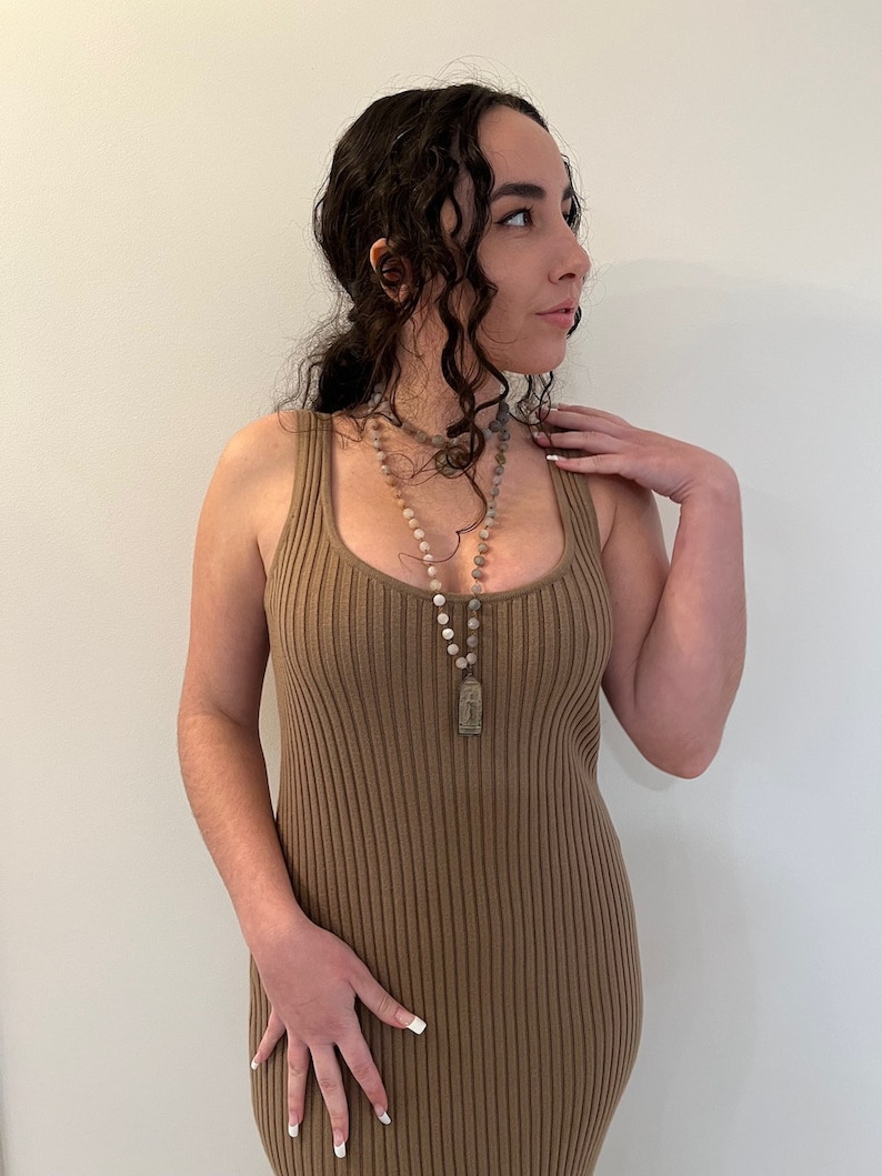 May include: A woman wearing a brown ribbed sleeveless dress with a layered beaded necklace and a pendant.