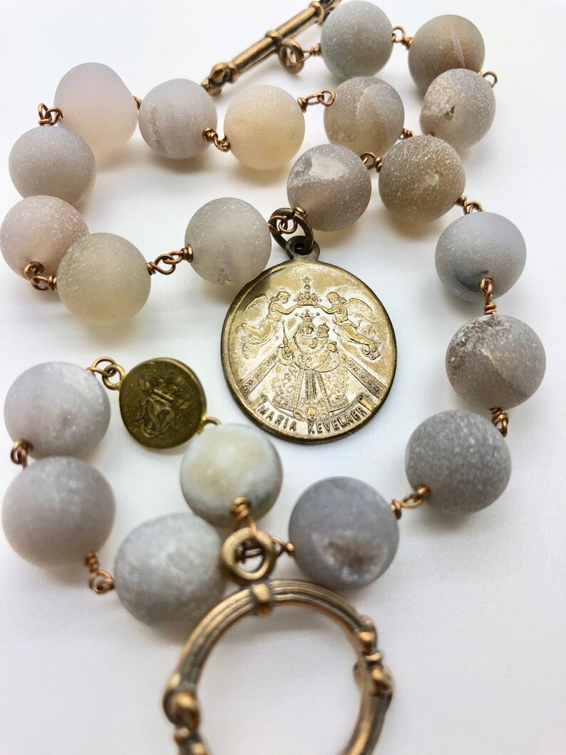 K&ouml;nnte beinhalten: Ein Rosenkranz mit gro&szlig;en, runden, grauen Perlen, die auf kupferfarbenem Draht aufgef&auml;delt sind. Ein kreisf&ouml;rmiger Anh&auml;nger mit den Worten "MARIA REVELAGE" ist angebracht. Der Rosenkranz hat einen Verschluss und zus&auml;tzliche dekorative Metallteile.