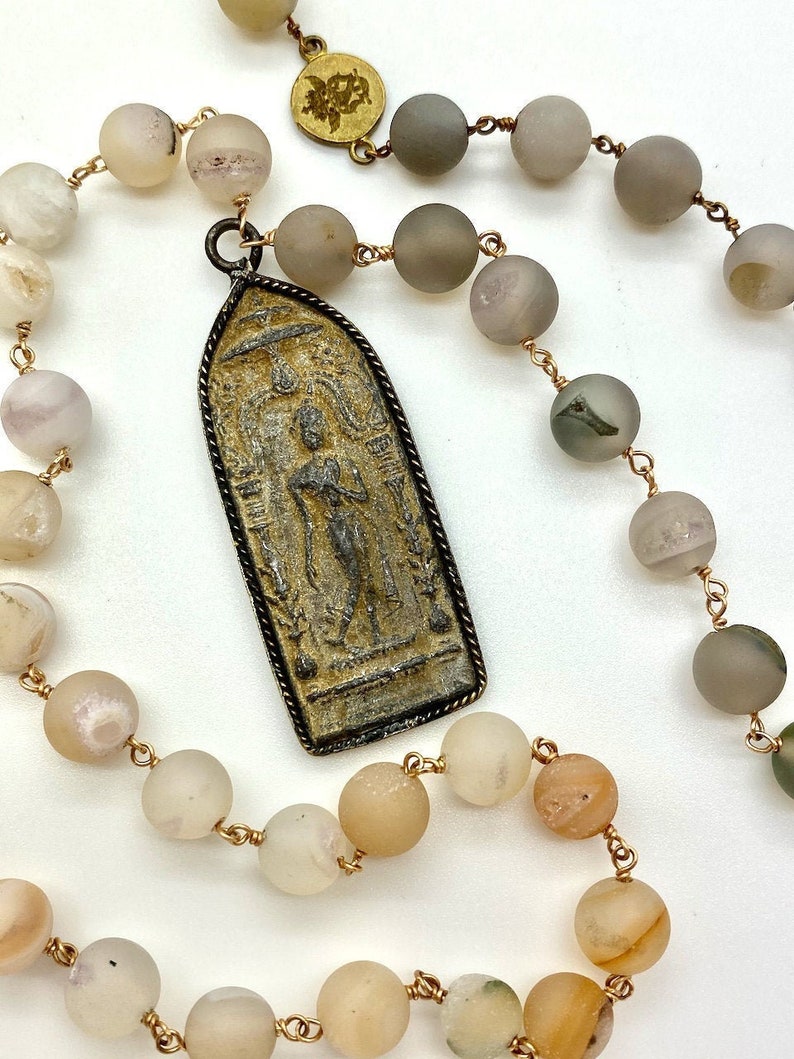 May include: A long necklace with a gold chain and a pendant featuring a detailed image of a person. The necklace is made with light brown and gray beads.