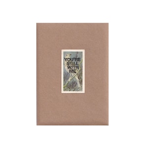 May include: A greeting card featuring a rectangular illustration of a bird on a branch, with the text "YOU'RE STILL WITH ME" in white. The card is set against a brown background.