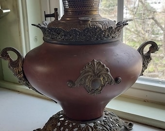 May include: Antique oil lamp with a burgundy body and ornate brass accents. The lamp features a decorative base, handles, and a detailed top with a mesh screen. It is a vintage piece, likely for home decor or a collector.