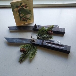 May include: Two antique wooden marking gauges with dark blades and wooden handles, arranged with a Christmas card and pine sprigs. The card reads "SEASON'S GREETINGS" and features a bell illustration. The gauges are positioned on a light-colored surface.