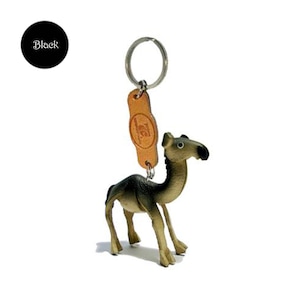 Op de afbeelding: Een kameel sleutelhanger met een metalen ring en een leren label. Het kameelbeeldje is bruin en donkergroen. Het label is bruin en heeft een logo. Een zwarte cirkel met het woord "Black" staat in de linkerbovenhoek.