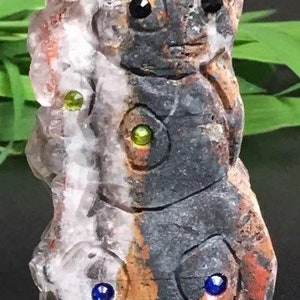 May include: A carved stone sculpture of three faces with green and blue eyes. The sculpture is made of a light gray and dark gray stone with brown and red accents.