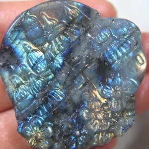 May include: A carved labradorite gemstone with a floral and architectural design. The stone displays iridescent blues, greens, and golds. The carved details include stylized flowers and building-like structures, creating a unique, artistic piece.