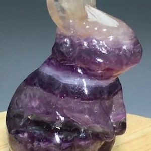May include: A carved rabbit figurine made of purple and white fluorite crystal. The bunny is in a sitting position with its ears upright. The crystal has bands of color throughout.